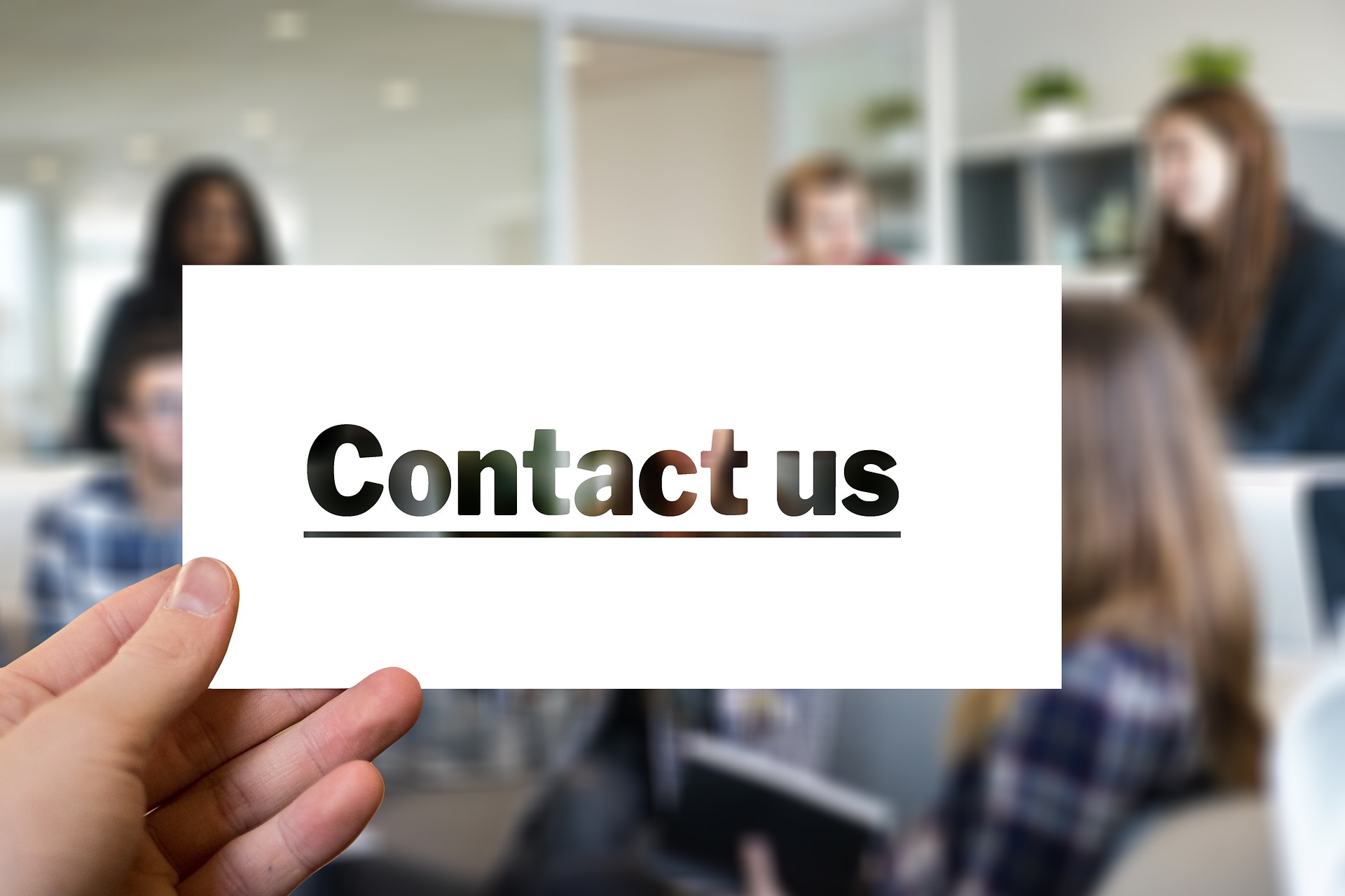 Hand holding a white card with bold, underlined text reading “Contact us.” The background is blurred, showing an office environment with several people.