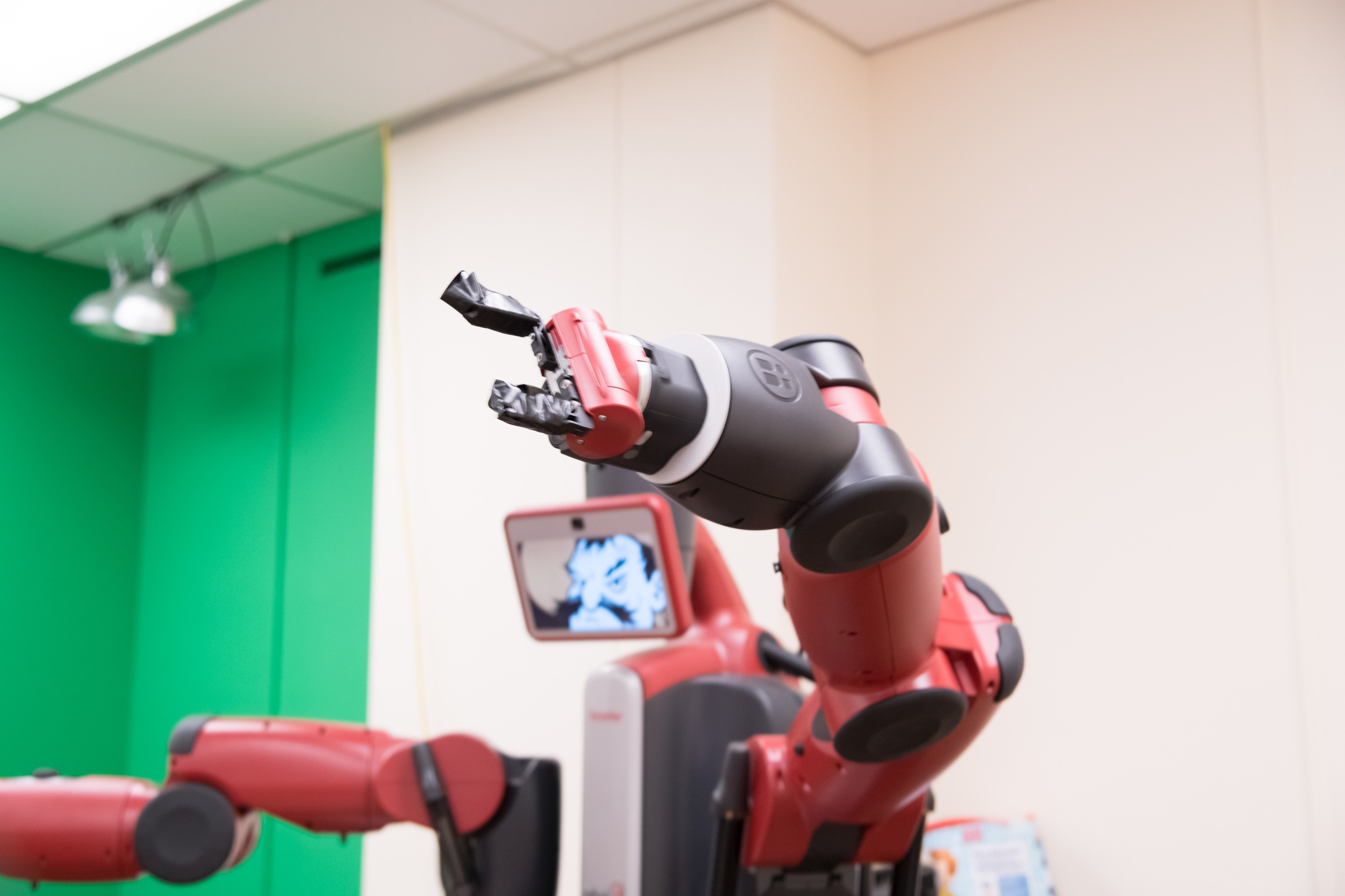 Red and black robotic arm with a gripper and a screen displaying a face-like graphic, set against green and beige walls