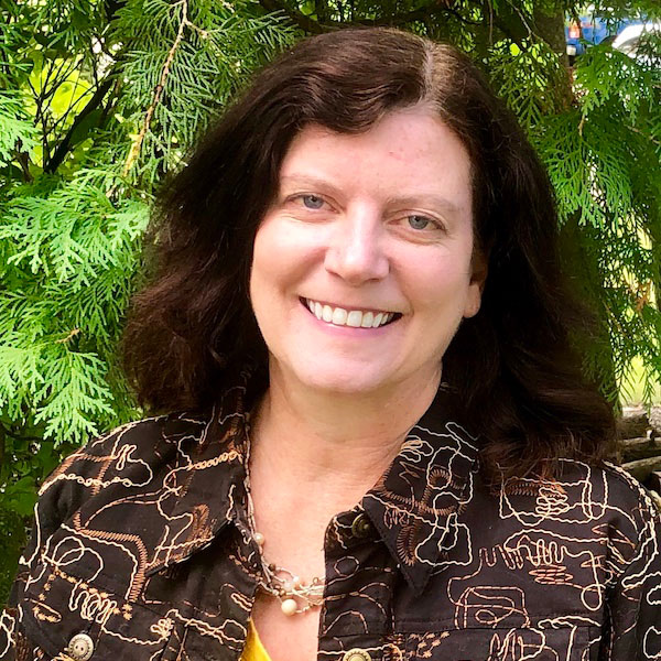 A woman wearing a brown, printed button-up shirt with a yellow shirt underneath smiles in front of a background of trees