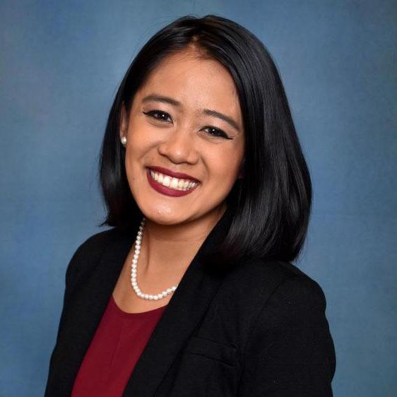 A woman with black hair wearing a red shirt, black blazer, and white pearl necklace smiles in front of a blue background