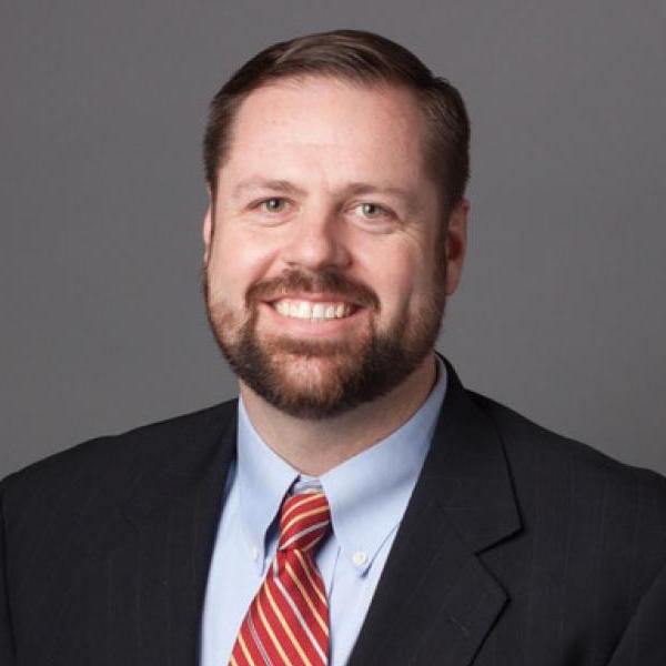A man with brown hair and a beard wearing a suit and site smiles in front of a gray background