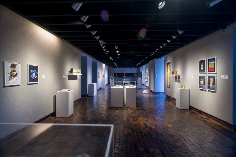 A glass case that contains historical items including a doll, shirt, and game. Interior of the Stillwater Museum of Art with dark brown wood floors, white walls. Colorful artwork hangs from the walls. Items are in white pedestals under display cases.