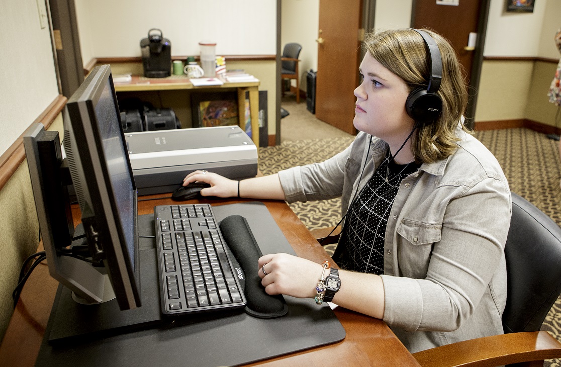 A female student with shoulder-length brown hair sits at a computer with headphones as she processes oral histories.