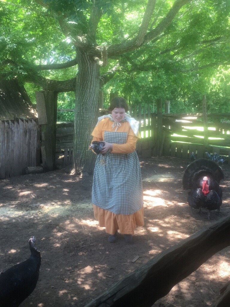 A female student with brown hair stands outside in 19th century rural period dress. A wild turkey stands next to her.