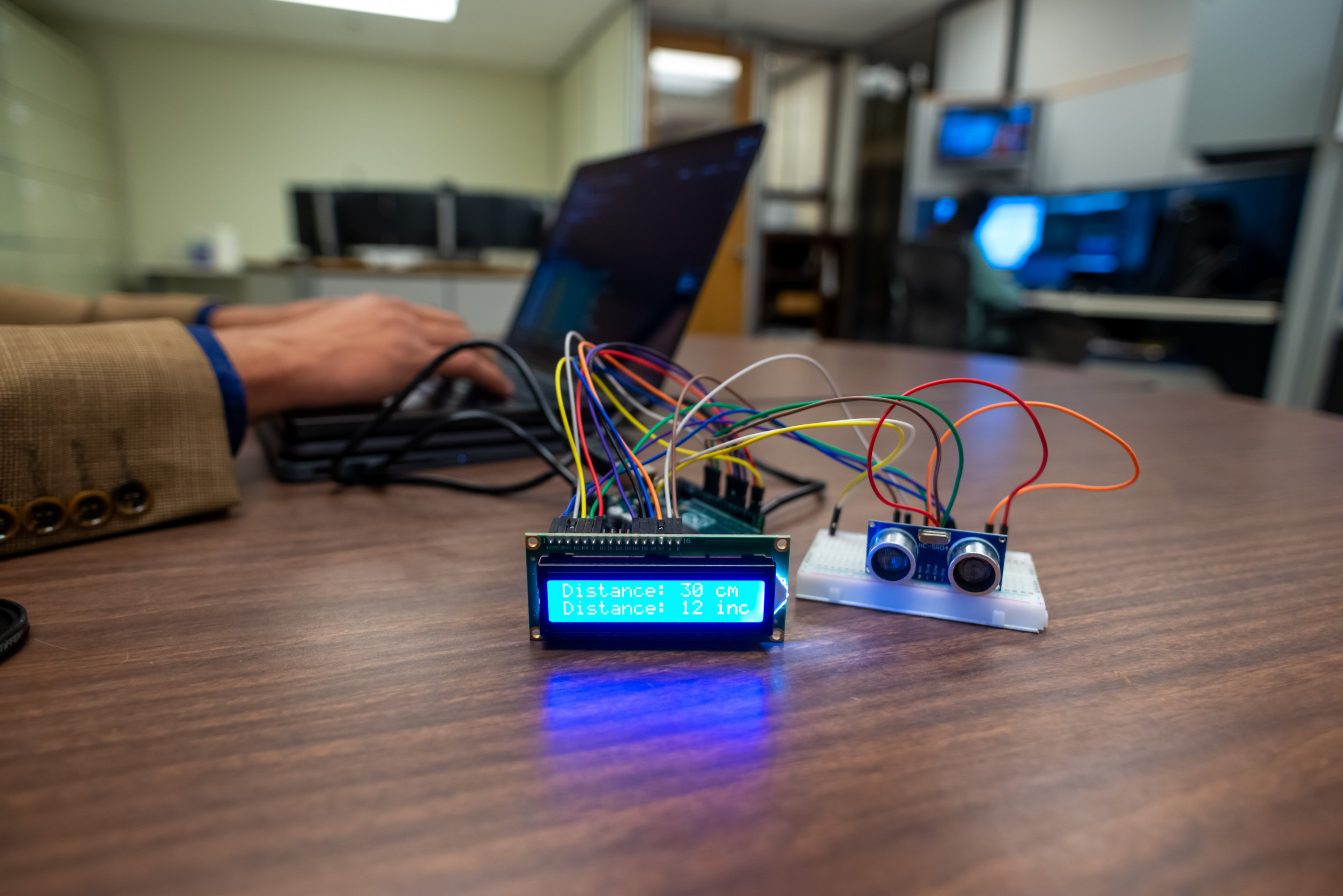 Person working on a laptop displaying code, with a breadboard and microcontroller circuit setup on a desk in an office environment