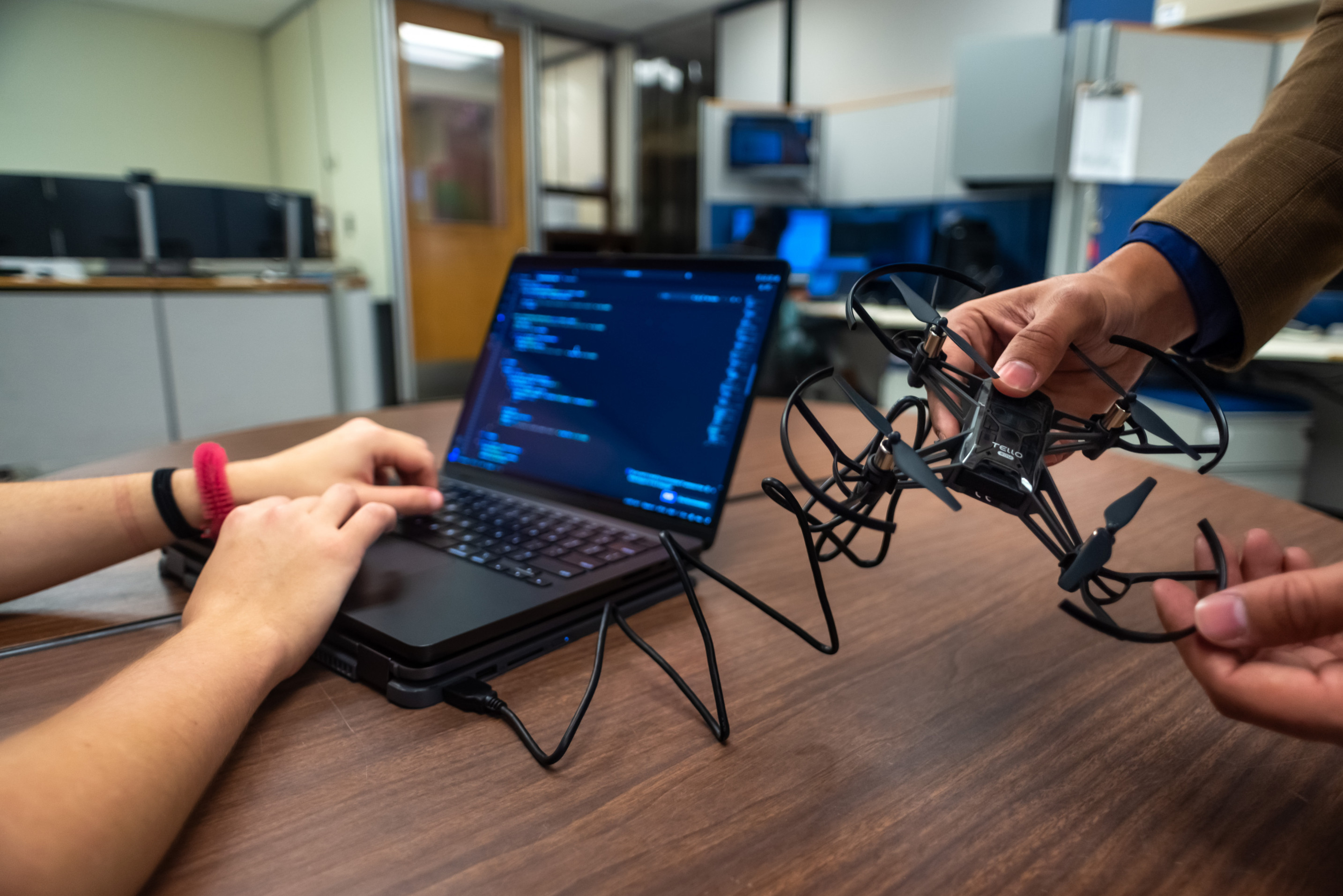 erson typing on a laptop displaying code, while another person holds a small drone beside it in an office or lab setting