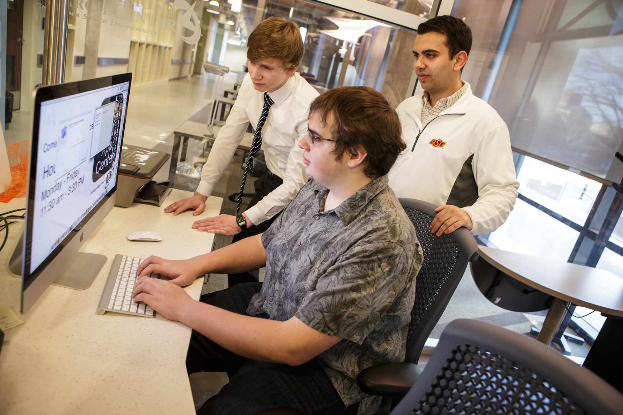 Three individuals in a modern office setting; one seated at a desk using a computer while two others stand nearby observing. Three individuals in a modern office setting; one seated at a desk using a computer while two others stand nearby observing.