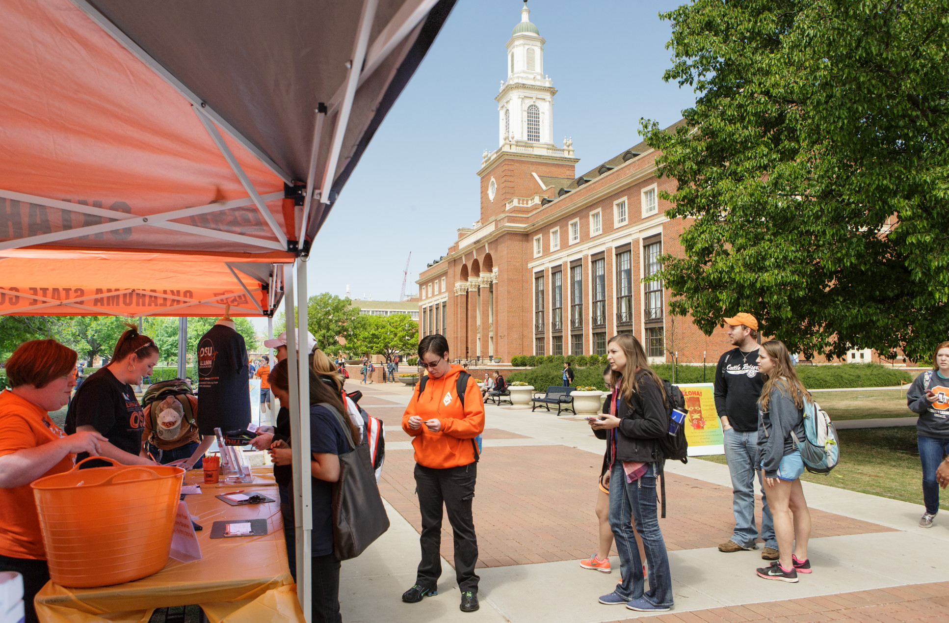 Group of people gathered outdoors on a college campus near a tent, with some interacting with staff or volunteers. A large brick building with white columns and a clock tower is visible in the background. Others are standing, using their phones, or chatting. Clear daytime weather. Group of people gathered outdoors on a college campus near a tent, with some interacting with staff or volunteers. A large brick building with white columns and a clock tower is visible in the background. Others are standing, using their phones, or chatting. Clear daytime weather.