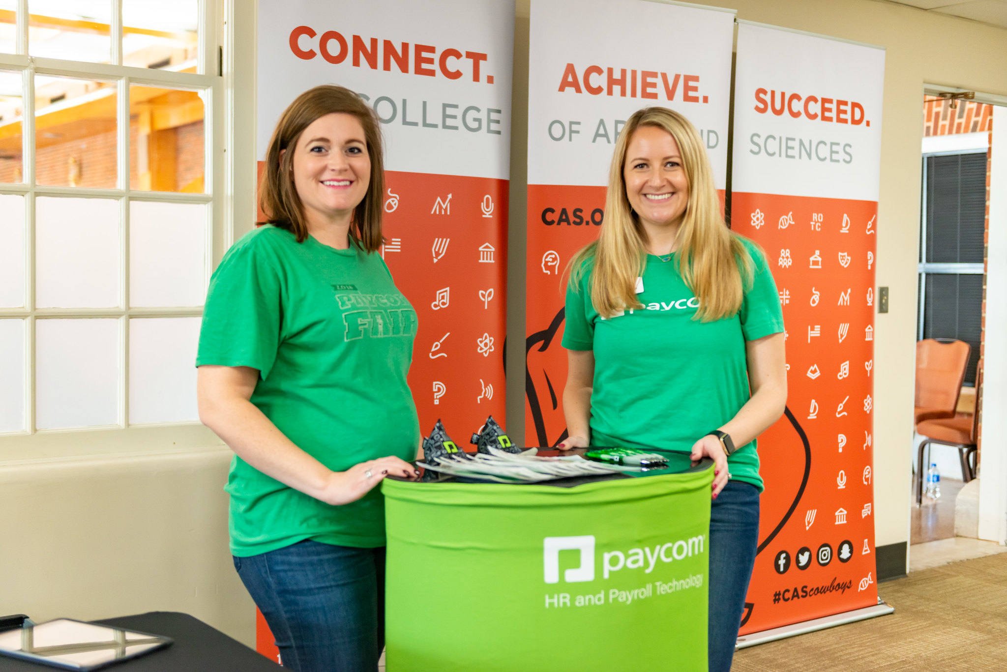Two individuals standing behind a green Paycom table with 