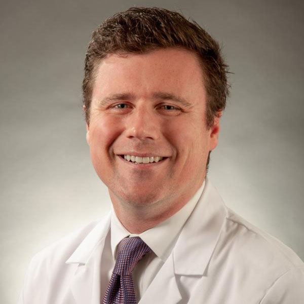 A man with brown hair and a white surgical coat smiles in front of an off-white background