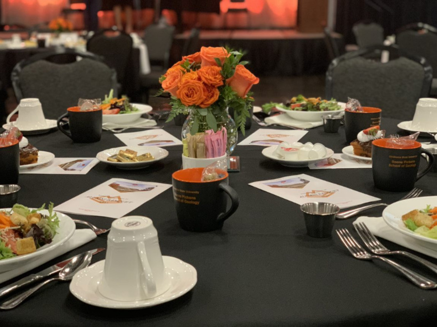 Banquet table set up with plates of salad and programs along with a orange roses as a center piece.