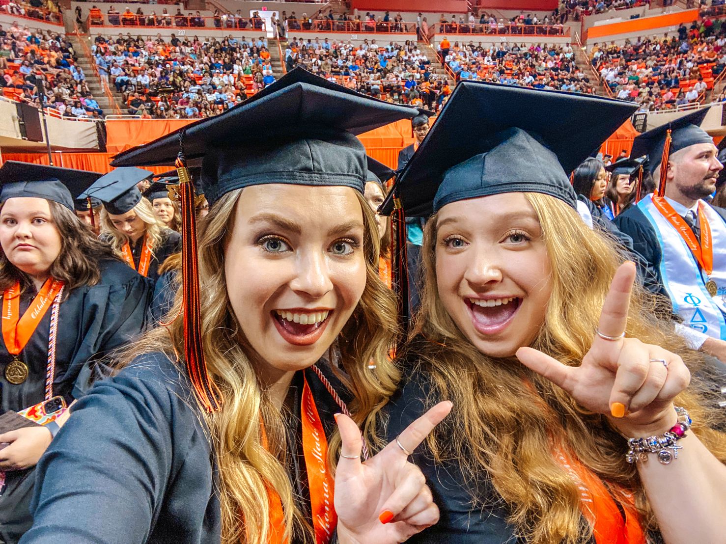 Students Megan and Izabelle making pistol pete hand sign at graduation in their caps and gowns.