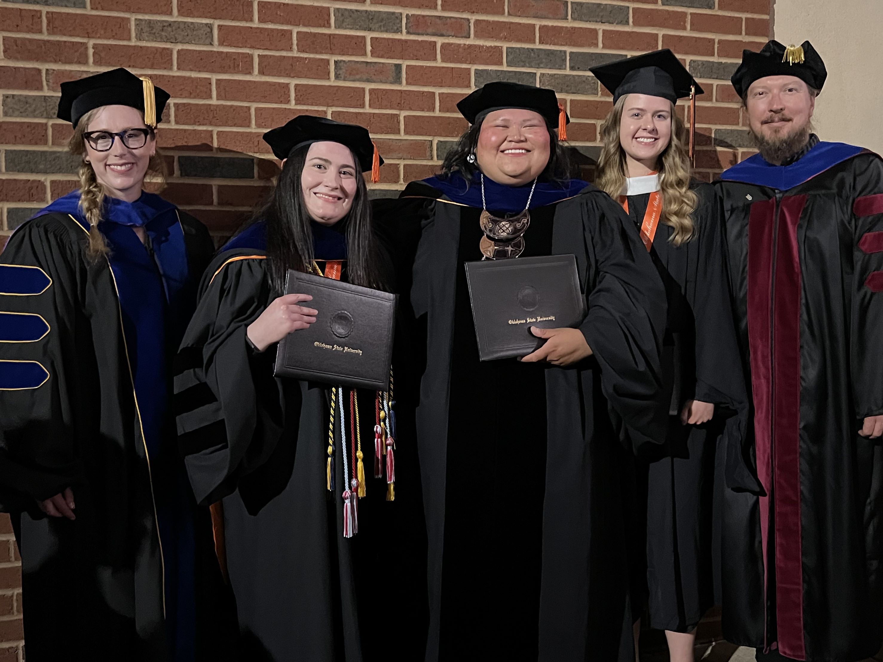 Five people standing against a wall in graduation attire.