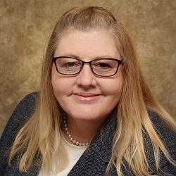A headshot of a woman with long blond hair, glasses, a white blouse and gray blazer with pearls.