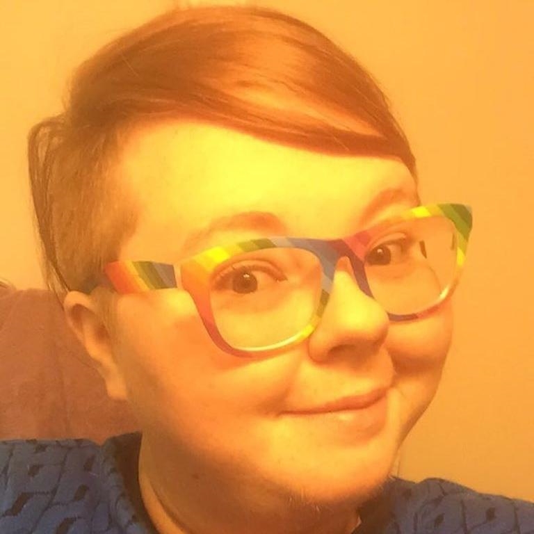 A man with brown hair wearing a blazer, tied, and beard stands outside next to a brick building.