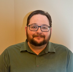 A man with brown hair, beard, and glasses in a green polo shirt stands in front of a neutral backdrop.