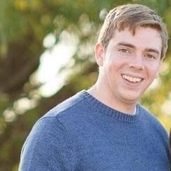 A man with brown hair and a blue shirt smiles with a natural background of greenery. A man with brown hair and a blue shirt smiles with a natural background of greenery.