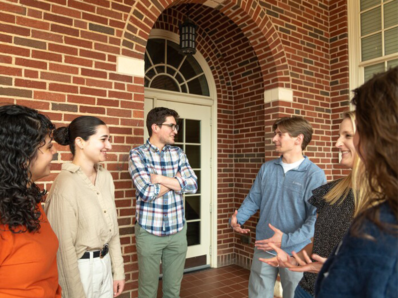 Graduate students standing outside in front of a door, engaged in discussion.