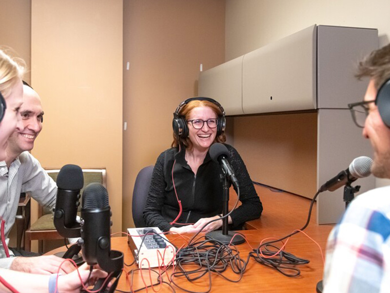 Four history experts/faculty sitting around a table with headphones and podcast equipment, recording a podcast and laughing.
