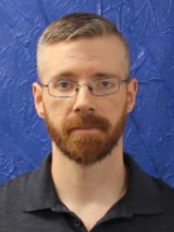 A man with short hair, a beard, and glasses wearing a black shirt against a blue background.