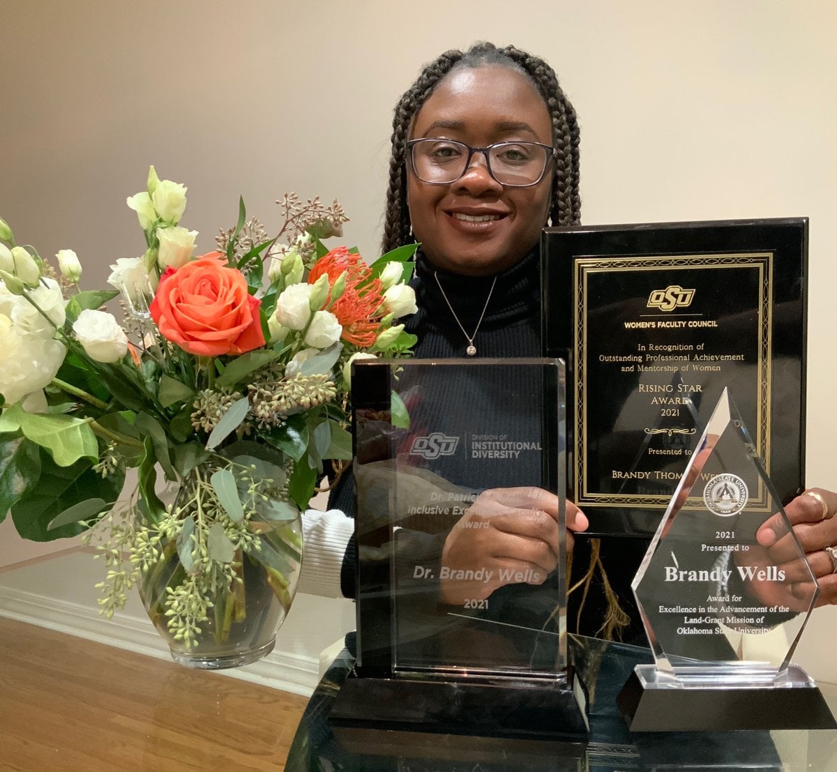 Dr. Wells, a Black woman wearing a black turtleneck and glasses stands next to her three awards and a flower arrangement.