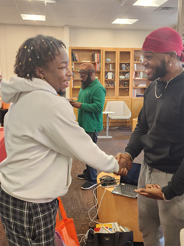 Two black male students greeting with a smile and hand shake while a center professor standing in the background