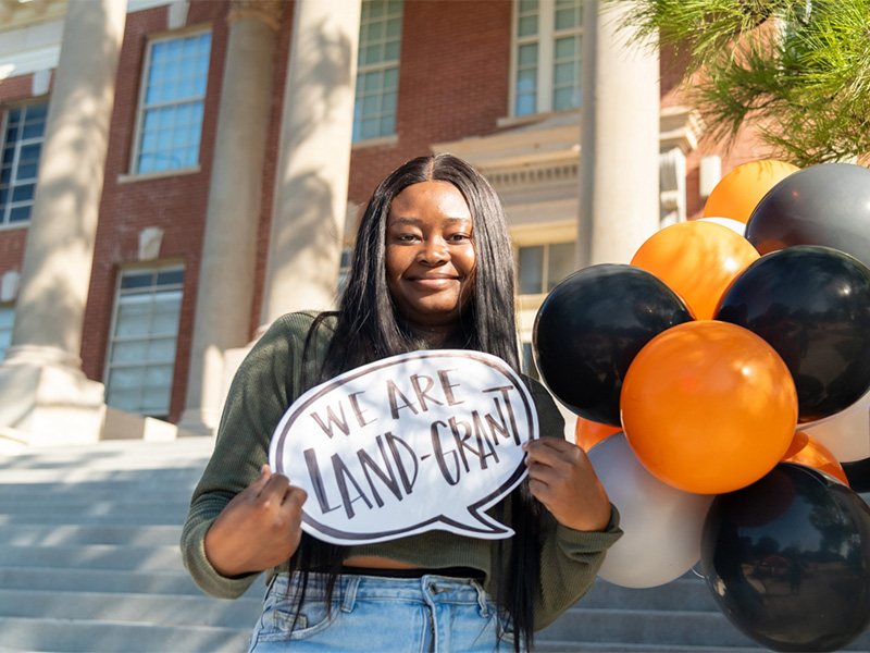A student holding a Land Grant sign, smiling and posing for the photo
