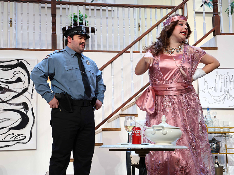 A theatrical stage scene featuring a police officer and a woman in a bright pink gown