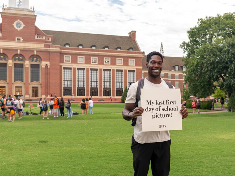A male student holding a sign that reads, 'This is my last first day of school' with a big smile, standing in front of a school building.